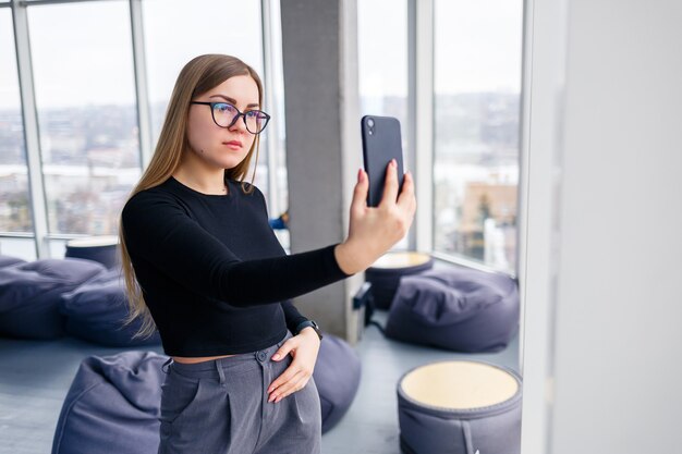 Donna d'affari adulta professionista impegnata a mandare SMS sul suo telefono cellulare indossando camicia e pantaloni in piedi davanti a grandi finestre di vetro.