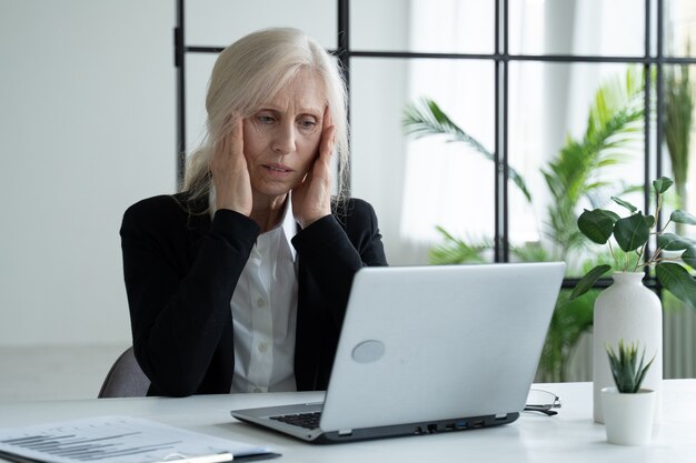 Donna d'affari anziana stanca che soffre di affaticamento degli occhi dopo aver lavorato su un laptop in ufficio