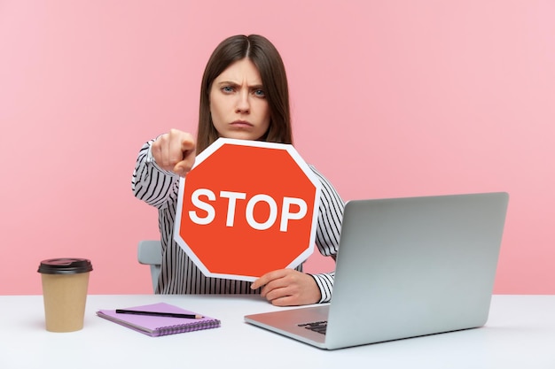 Donna d'affari arrabbiata che mostra il simbolo di arresto che punta il dito verso la fotocamera, mostrando il segnale stradale rosso seduto sul posto di lavoro, concetto di divieto negativo. Studio indoor girato isolato su sfondo rosa