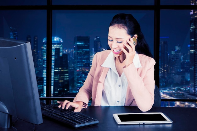 Donna d'affari asiatica che fa gli straordinari al telefono in ufficio con gli edifici per uffici del paesaggio urbano
