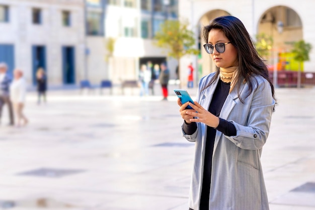 Donna d'affari asiatica che indossa un abito sms sullo smartphone in piedi in strada in una giornata di sole