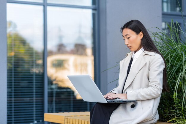 Donna d'affari asiatica che lavora con il laptop fuori dall'ufficio, seduta su una panchina durante la pausa di lavoro