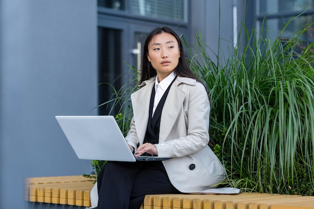 Donna d'affari asiatica che lavora con il laptop fuori dall'ufficio, seduta su una panchina durante la pausa di lavoro