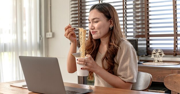 Donna d'affari asiatica che lavora da sola online a casa. Donna che mangia la tagliatella della tazza e occupata.