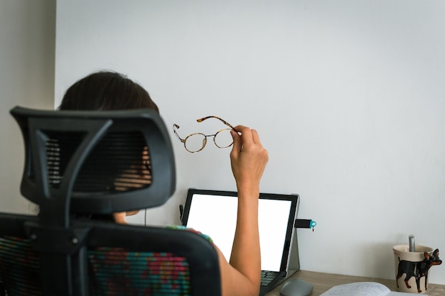 Donna d'affari asiatica che tiene gli occhiali mentre fa gli straordinari sul computer portatile a casa.