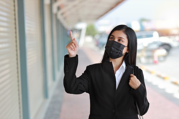 Donna d'affari asiatica con maschera protettiva per camminare sulla strada pubblica all'aperto e puntare qualcosa.