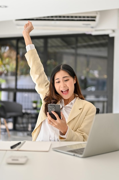 Donna d'affari asiatica felicissima alla sua scrivania che guarda lo schermo del suo telefono alzando la mano