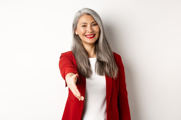 Donna d'affari asiatica professionale con i capelli grigi, salutando, allungando la mano per la stretta di mano e sorridendo, in piedi su sfondo bianco