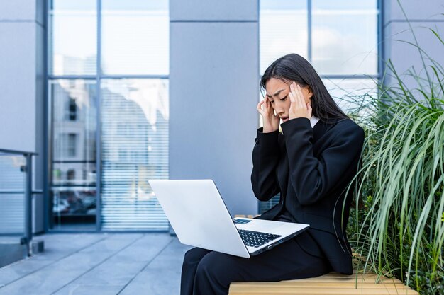 Donna d'affari asiatica sconvolta e confusa che lavora al computer portatile, fuori dall'ufficio, seduta su una panchina