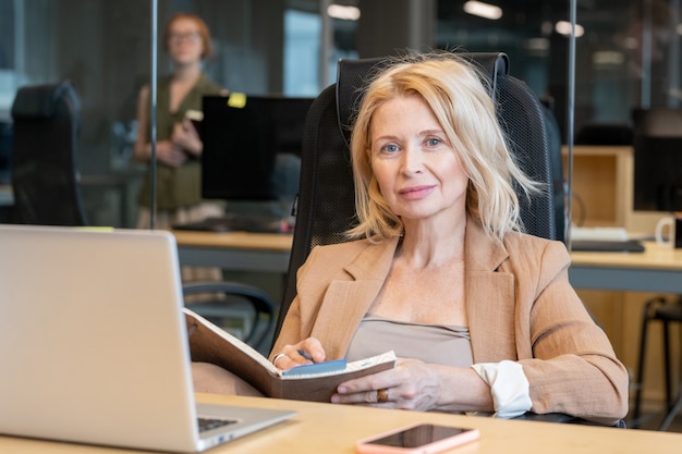 Donna d'affari bionda matura contemporanea con taccuino aperto che prende appunti sugli appuntamenti o pianifica il lavoro mentre è seduto alla scrivania