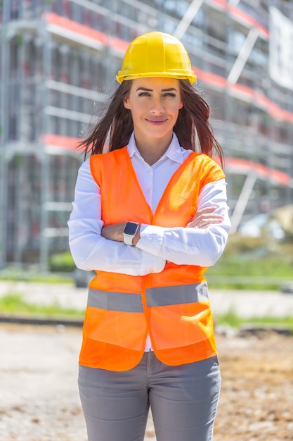 Donna d'affari che indossa jeans, giubbotto di sicurezza e casco sta con sicurezza di fronte alla costruzione coperta da impalcature.