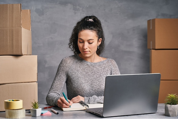 Donna d'affari che lavora da casa con laptop e blocco note che prepara scatole con prodotti per l'invio