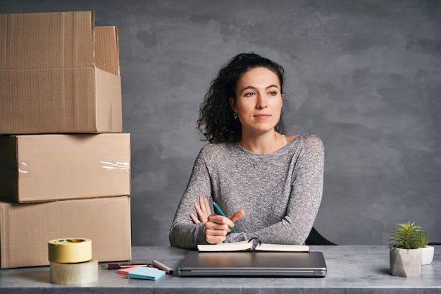 Donna d'affari che lavora da casa con laptop e blocco note che prepara scatole con prodotti per la consegna