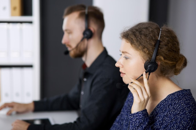 Donna d'affari che parla con l'auricolare mentre è seduto con un collega dalla barba rossa in un ufficio moderno. Telemarketing e servizio clienti. Gruppo di persone diverse nel call center.