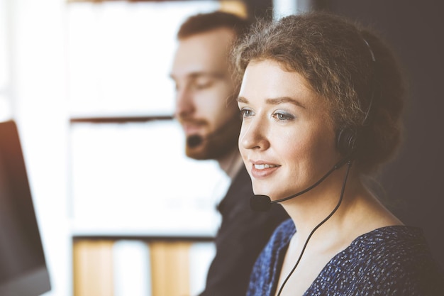 Donna d'affari che parla in cuffia mentre è seduto con un collega dalla barba rossa in un ufficio soleggiato. Gruppo di persone diverse nel call center.