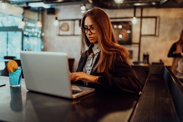 Donna d'affari con gli occhiali seduta in un caffè moderno e che lavora sul suo laptop mentre beve un cocktail Messa a fuoco selettiva