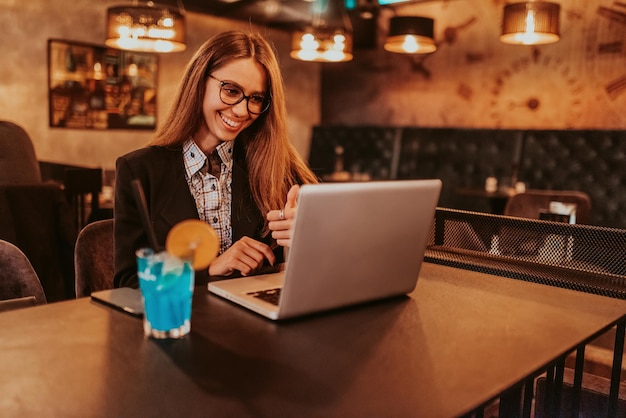 Donna d'affari con gli occhiali seduta in un caffè moderno e che lavora sul suo laptop mentre beve un cocktail Messa a fuoco selettiva