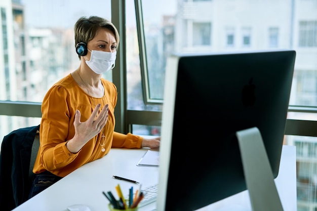 Donna d'affari con maschera facciale che lavora su PC desktop e comunica con qualcuno tramite l'auricolare in ufficio