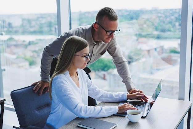 Donna d'affari con un laptop per un collega di sesso maschile che guarda in un taccuino aperto Collaborazione aziendale e lavoro di squadra Millennial europei al tavolo in ufficio Persone moderne di successo