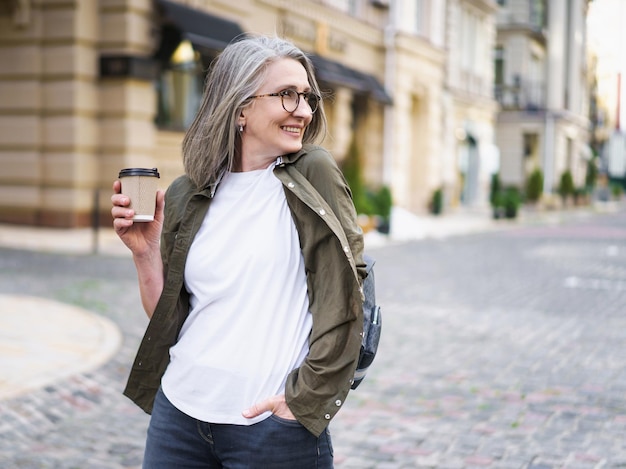 Donna d'affari con una tazza usa e getta con caffè caldo Donna matura che trascorre il tempo libero viaggiando nella città vecchia europea gustando una tazza di tè e caffè Donna dai capelli grigi in casual con zaino