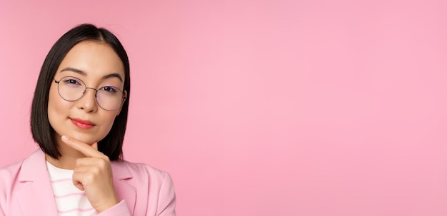 Donna d'affari coreana che pensa con gli occhiali che guarda premurosa alla macchina fotografica che prende una decisione in piedi su sfondo rosa