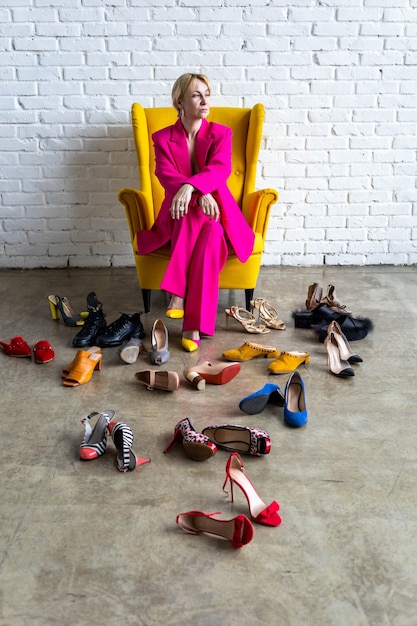 Donna d'affari di moda in abito rosa brillante seduto su una poltrona gialla nella stanza del soppalco casualmente molte scarpe