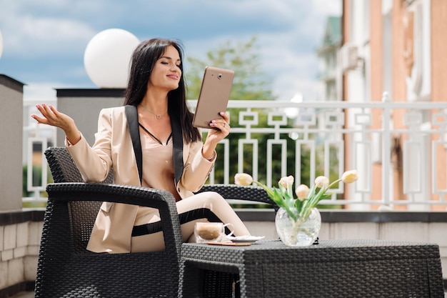 Donna d'affari di successo in giacca e cravatta che parla in videoconferenza utilizzando un tablet fuori dalla terrazza dell'hotel