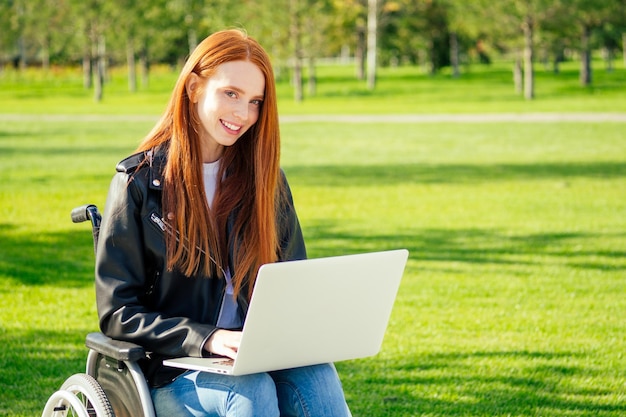 Donna d'affari di zenzero dai capelli rossi che si sente felice, lei seduta su sedia a rotelle con il laptop, indossa una calda giacca di pelle nel parco estivo.