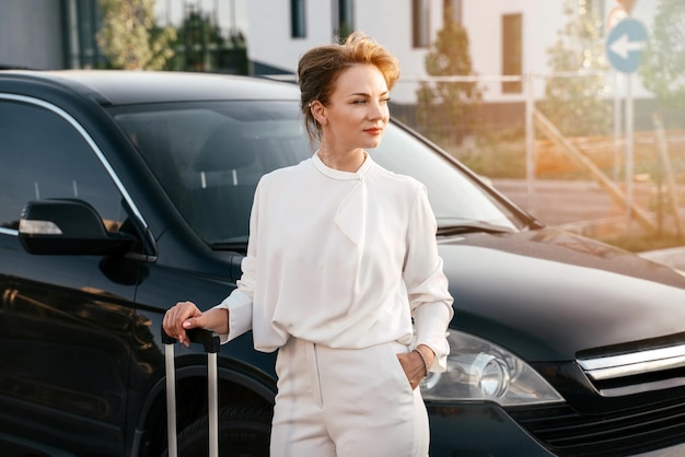 Donna d'affari elegante di successo con borsa da viaggio in piedi vicino all'auto, distogliendo lo sguardo. Viaggio di lavoro