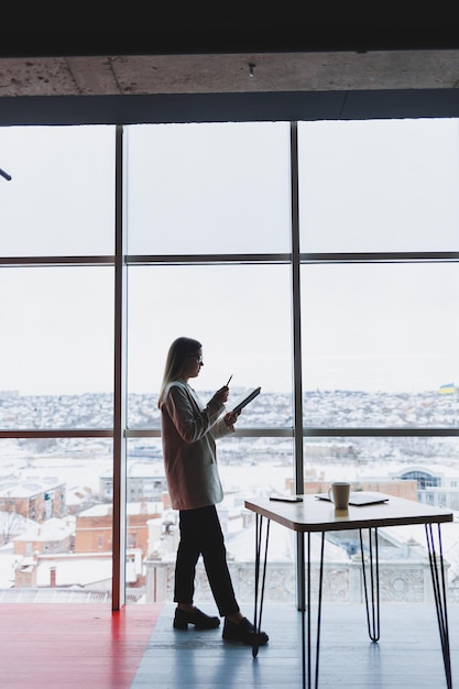 Donna d'affari esperta che tiene un telefono mentre si trova in un moderno ufficio interno vicino a una grande finestra che si affaccia sul paesaggio urbano Il dirigente femminile sembra soddisfatto dopo un incontro di successo