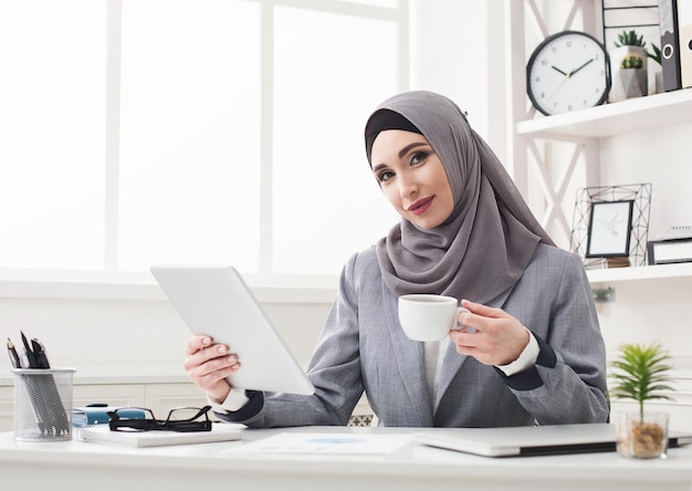 Donna d'affari in hijab che lavora in ufficio, usa tablet e beve caffè