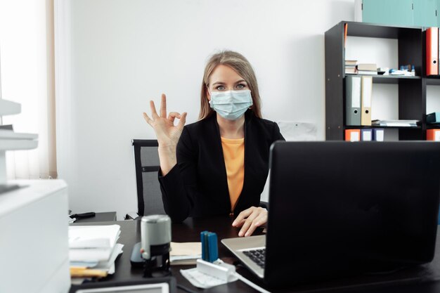 Donna d'affari in maschera medica che lavora su un laptop nell'area di lavoro in ufficio