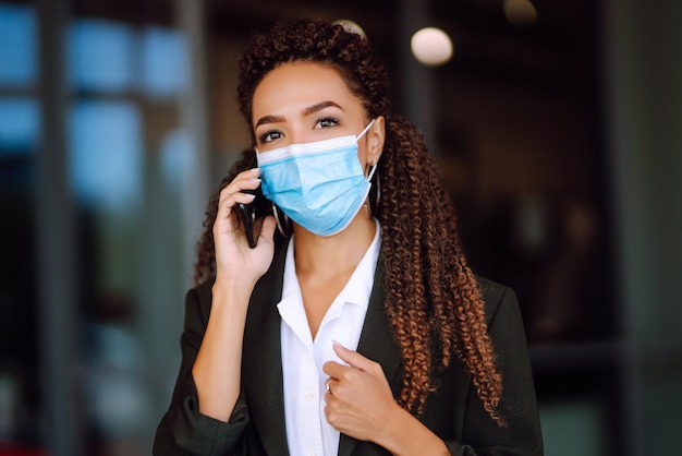 Donna d'affari in maschera medica protettiva in piedi vicino all'ufficio e usa il telefono covid19