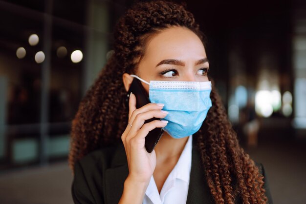 Donna d'affari in maschera medica protettiva in piedi vicino all'ufficio e usa il telefono covid19