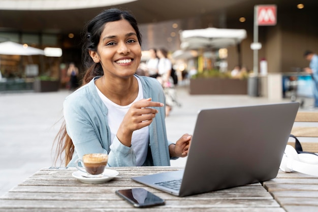 Donna d'affari indiana sorridente che utilizza un computer portatile che lavora online a bere caffè nella caffetteria