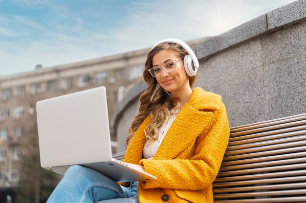 Donna d'affari indossare occhiali ascoltare musica cuffia seduta all'aperto panca utilizzando laptop all'aperto vestito elegante cappotto giallo sorriso femmina caucasica anni '30 Goditi podcast o audiolibri all'esterno