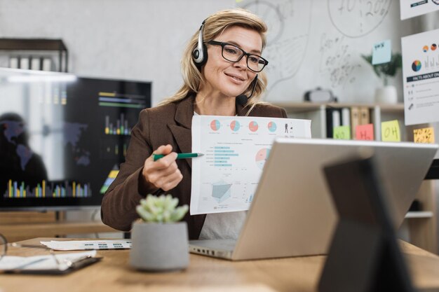 Donna d'affari invecchiata caucasica che utilizza cuffie e laptop moderni per la videoconferenza in ufficio