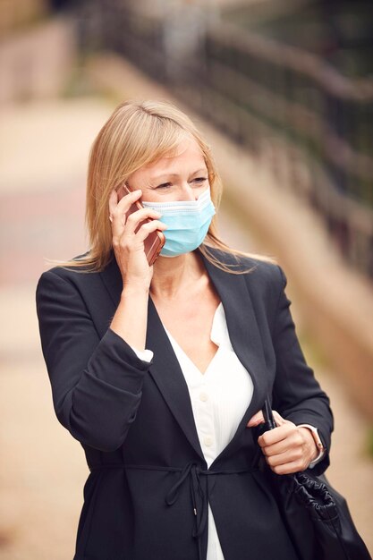 Donna d'affari matura con telefono cellulare che indossa una maschera facciale DPI cammina in strada durante la pandemia di salute