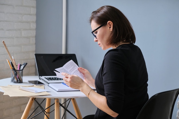 Donna d'affari matura seduta alla scrivania in ufficio a leggere documenti cartacei, corrispondenza commerciale, buste sul tavolo