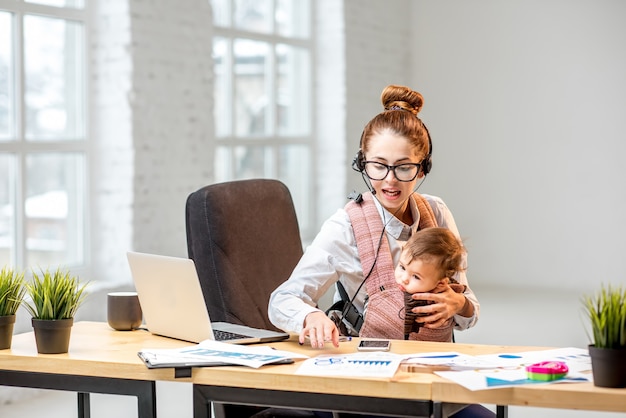 Donna d'affari multitasking che lavora con cuffie e laptop seduta con suo figlio bambino all'interno dell'ufficio bianco