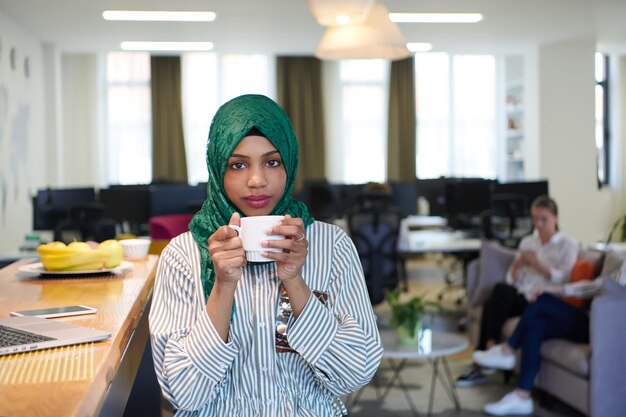 donna d'affari musulmana africana che indossa un hijab verde che beve tè mentre lavora su un computer portatile nell'area relax del moderno ufficio di avvio a pianta aperta. Diversità, concetto multirazziale