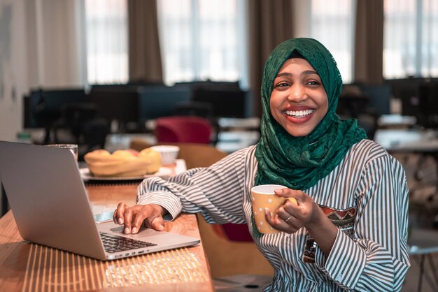 Donna d'affari musulmana africana che indossa un hijab verde che beve tè mentre lavora su un computer portatile nell'area relax del moderno ufficio di avvio a pianta aperta. Foto di alta qualità