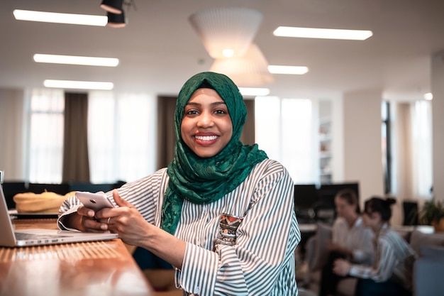 Donna d'affari musulmana africana che indossa un hijab verde utilizzando lo smartphone mentre si lavora su un computer portatile nell'area relax del moderno ufficio di avvio a pianta aperta. Foto di alta qualità