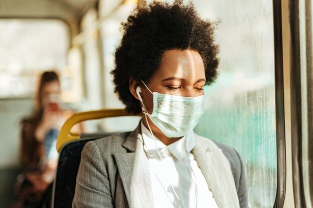 Donna d'affari nera con maschera facciale che ascolta musica con gli auricolari mentre si reca al lavoro in autobus durante l'epidemia di virus