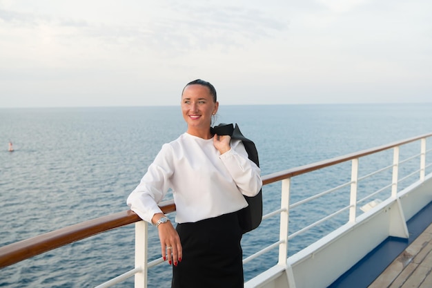 Donna d'affari o ragazza carina felice con la faccia sorridente in giacca grigia, ha la manicure rossa sul ponte della nave che guarda il mare, l'acqua dell'oceano con la barca a vela sullo sfondo del cielo nuvoloso, i viaggi e il turismo