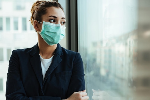 Donna d'affari pensierosa che indossa una maschera protettiva mentre è in piedi vicino alla finestra e guarda attraverso di essa