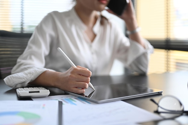 Donna d'affari ritagliata che ha una conversazione sul cellulare e utilizza la tavoletta digitale sul posto di lavoro