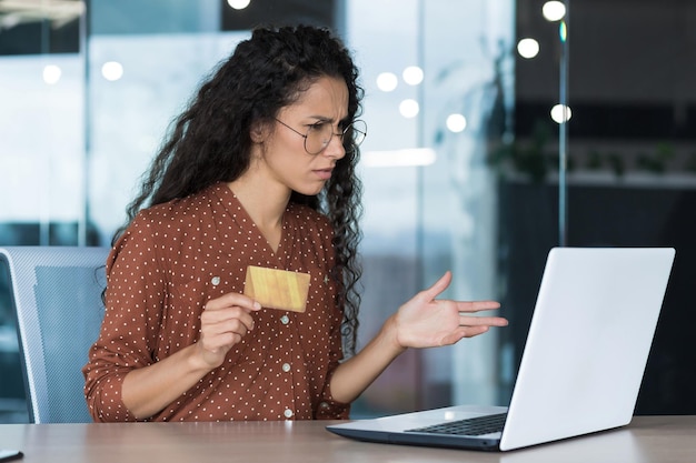 Donna d'affari sconvolta e delusa che lavora in un ufficio moderno utilizzando laptop e carta di credito bancaria