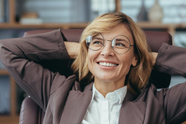 Donna d'affari senior bionda di successo con gli occhiali che si rilassa e sorride nel suo elegante ufficio a casa