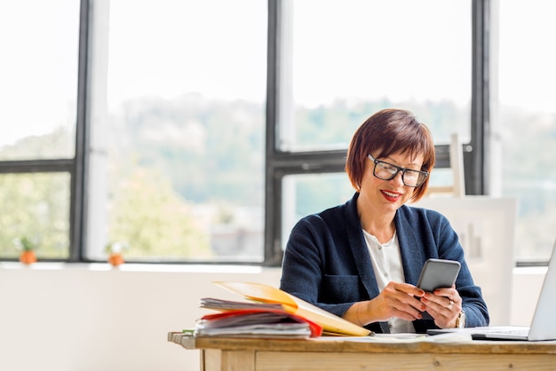 Donna d'affari senior che lavora con documenti e smartphone all'interno luminoso e moderno dell'ufficio